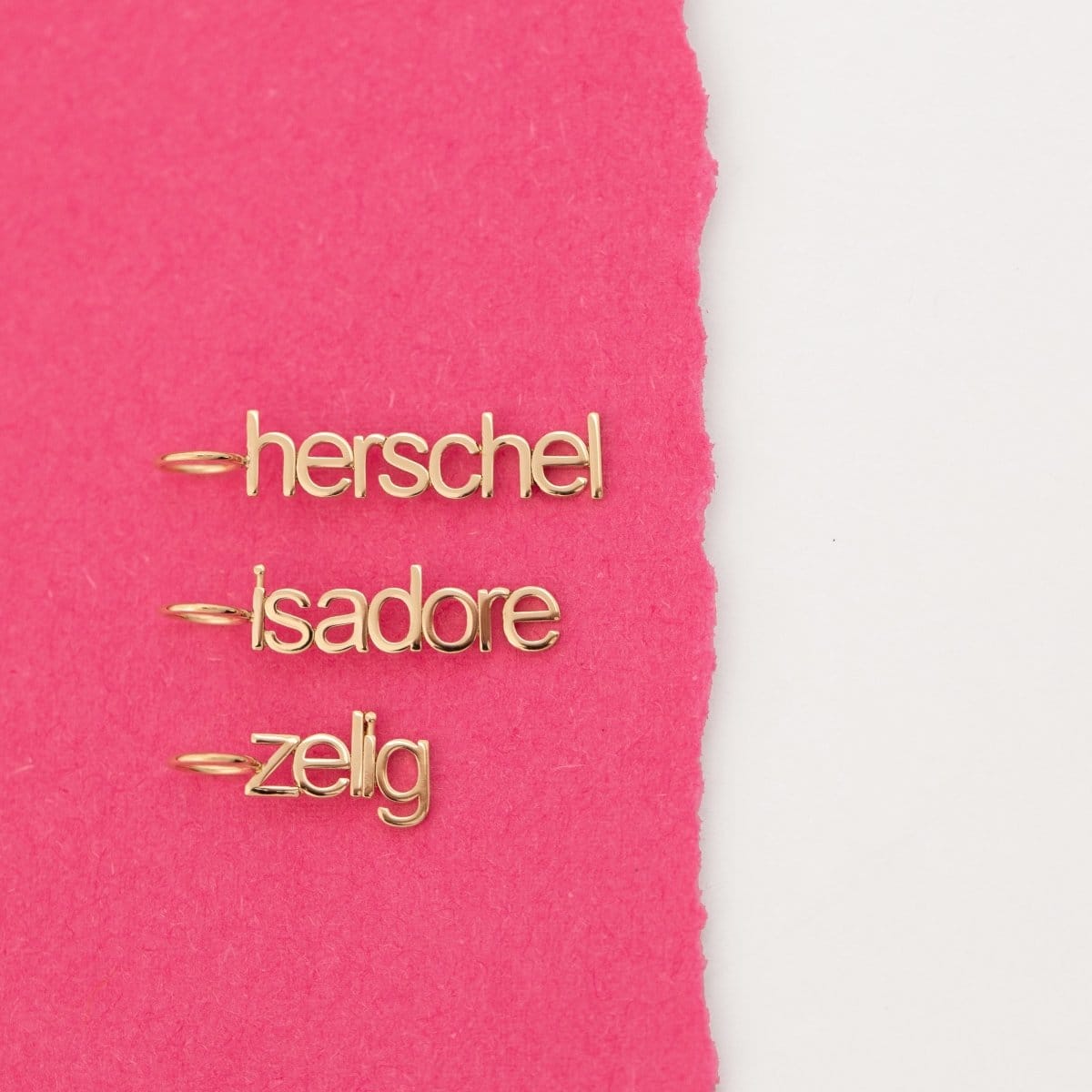 Three yellow gold Petite Nameplate Pendants with 'herschel', 'isadore', and 'zelig' on a pink and white background. 