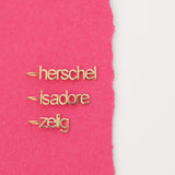 Three yellow gold Petite Nameplate Pendants with 'herschel', 'isadore', and 'zelig' on a pink and white background. 