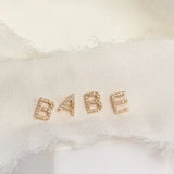 Gold 'BABE' letter pave earrings on a textured neutral background. 
