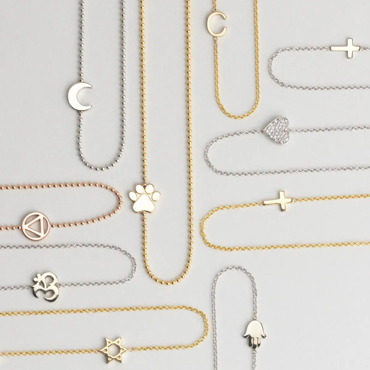 Several 14k Gold Asymmetrical Charm Necklaces in yellow, white and rose gold in both bead and cable chains (moon, paw, recovery, om, star of david, C, cross x 2 and a pavé heart) on a light grey background. 