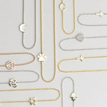 Several 14k Gold Asymmetrical Charm Necklaces in yellow, white and rose gold in both bead and cable chains (moon, paw, recovery, om, star of david, C, cross x 2 and a pavé heart) on a light grey background. 