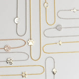 Several 14k Gold Asymmetrical Charm Necklaces in yellow, white and rose gold in both bead and cable chains (moon, paw, recovery, om, star of david, C, cross x 2 and a pavé heart) on a light grey background. 