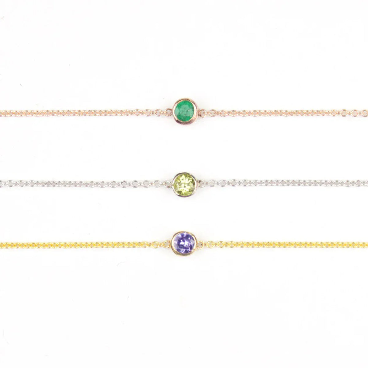 Three 14K Gold Asymmetrical Birthstone Necklaces - Emerald (May) in rose gold, Peridot (August) in white gold and Tanzanite (December) in yellow gold on a white background. 