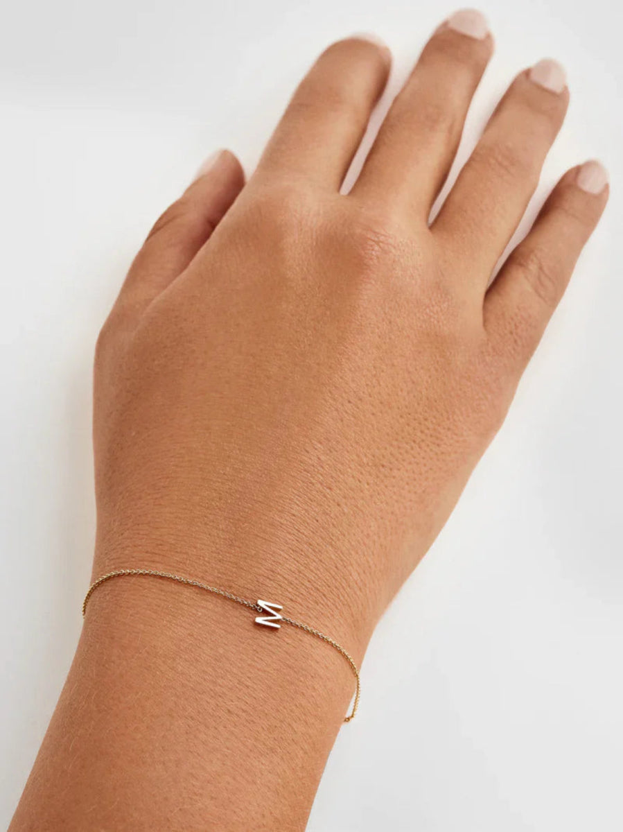 Hand wearing a gold bracelet with an M charm a plain background. 