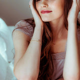 Gold bracelet with an M letter charm shown on a woman wearing a pink satin headband and lace lingerie, sitting on a bed. 