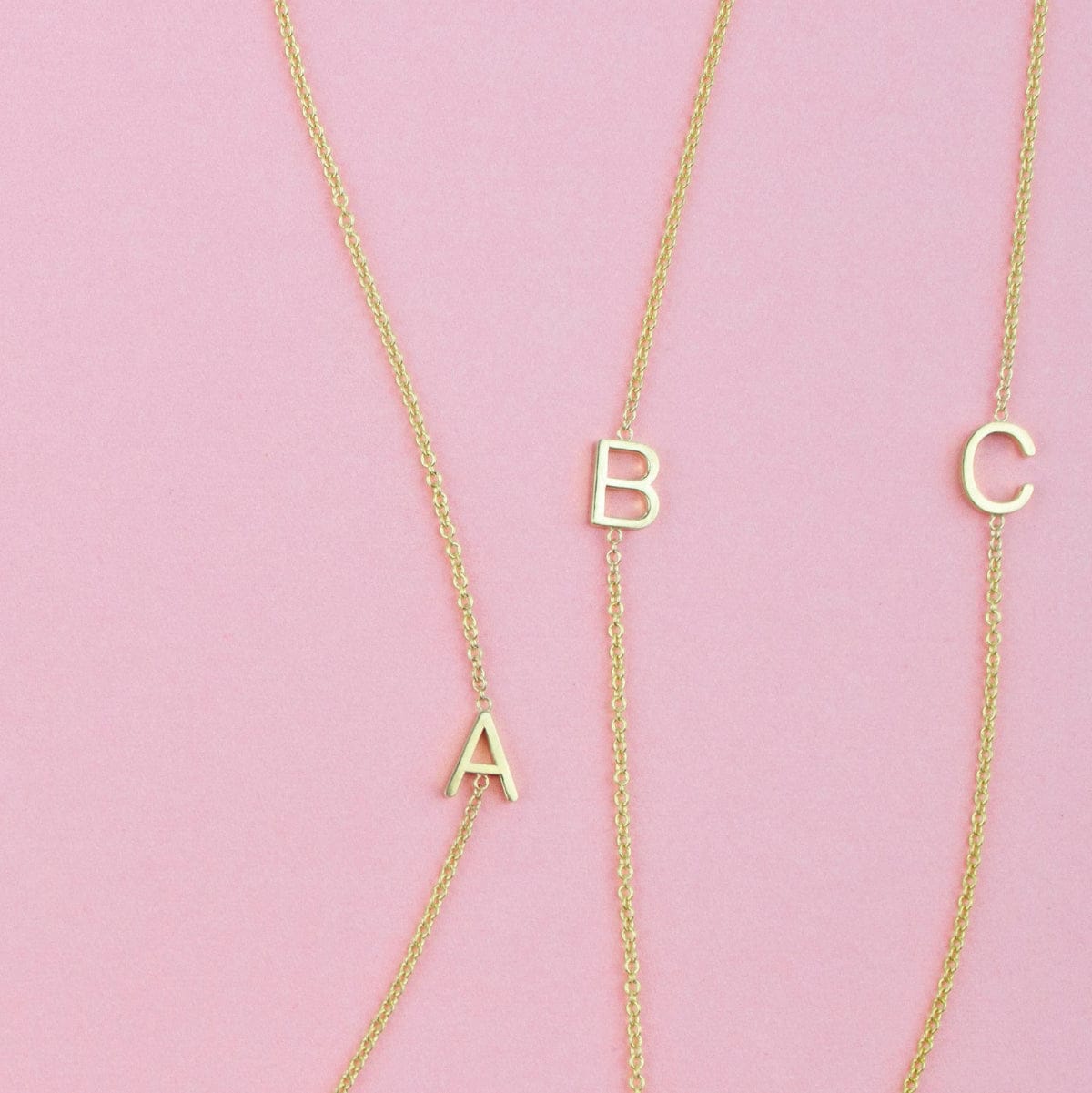 Three gold necklaces with single letter charms A, B and C on a pink background. 