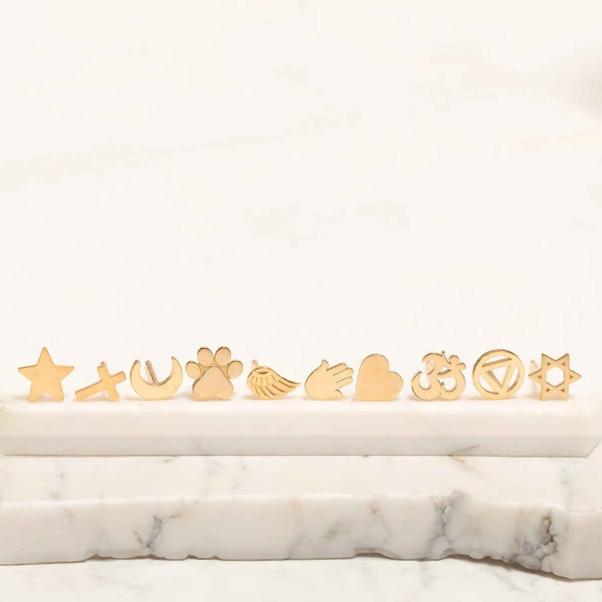 Several Charm Studs – Star, Cross, Moon, Paw, Angel Wing, Hamsa, Heart, Om, Recovery and Star of David laid on a marble stone surface and a white background. 