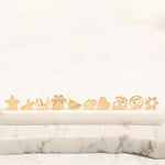 Several Charm Studs – Star, Cross, Moon, Paw, Angel Wing, Hamsa, Heart, Om, Recovery and Star of David laid on a marble stone surface and a white background. 