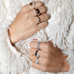 Hands wearing a Love on the Rocks Ring, two Make It a Double Rings, the Evermore Ring, two Cosmo Rings, a Martini Ring and  a Pinky Promise Ring, with a cream textured background. 