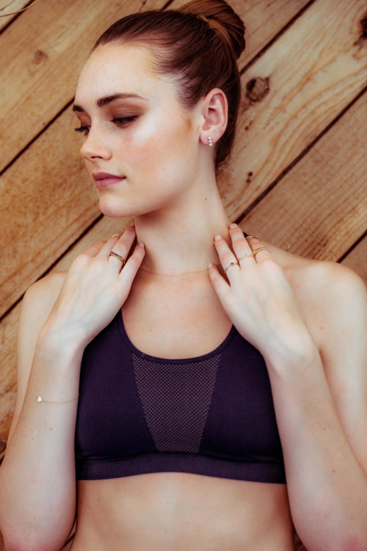 A woman wearing a yellow gold Custom Gold Bracelet with a heart charm, several gold rings, a necklace and earrings, in a dark purple sports bra against a wooden background.