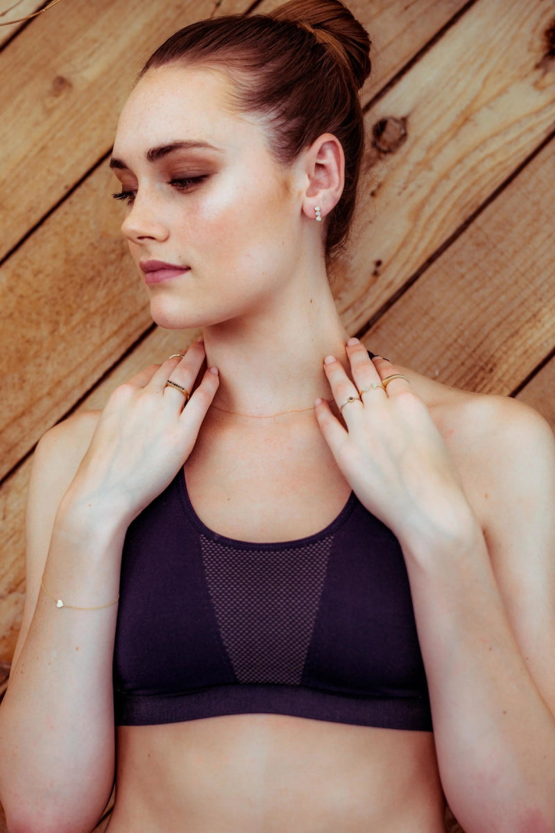 Gold heart charm bracelet shown on a woman wearing a dark purple sports bra against a wooden background. 