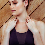 Gold heart charm bracelet shown on a woman wearing a dark purple sports bra against a wooden background. 