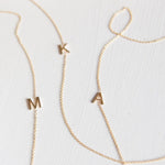 Three gold necklaces with single letter charms, being M, K and A on a plain background. 