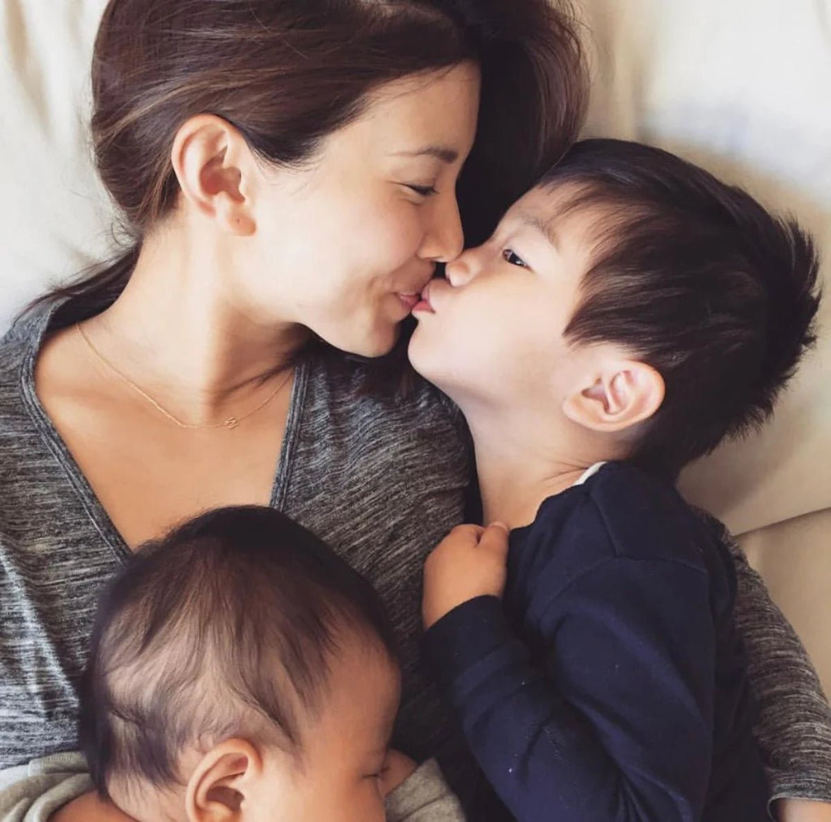 Woman holding two children close together and wearing a gold necklace with a letter M charm. 