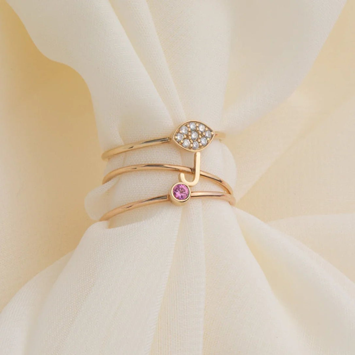 Three gold rings, including a yellow gold Pavé Petal Ring, a rose gold J 14k gold Letter Ring, and a rose gold Birthstone Ring on a cream fabric background.