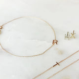 A Custom Gold Bracelet - 1 Letter with the letter R, two 14k Gold Letter Studs in H and X and a Custom Gold Necklace - 1 Letter with the letter T against a light marble background. 