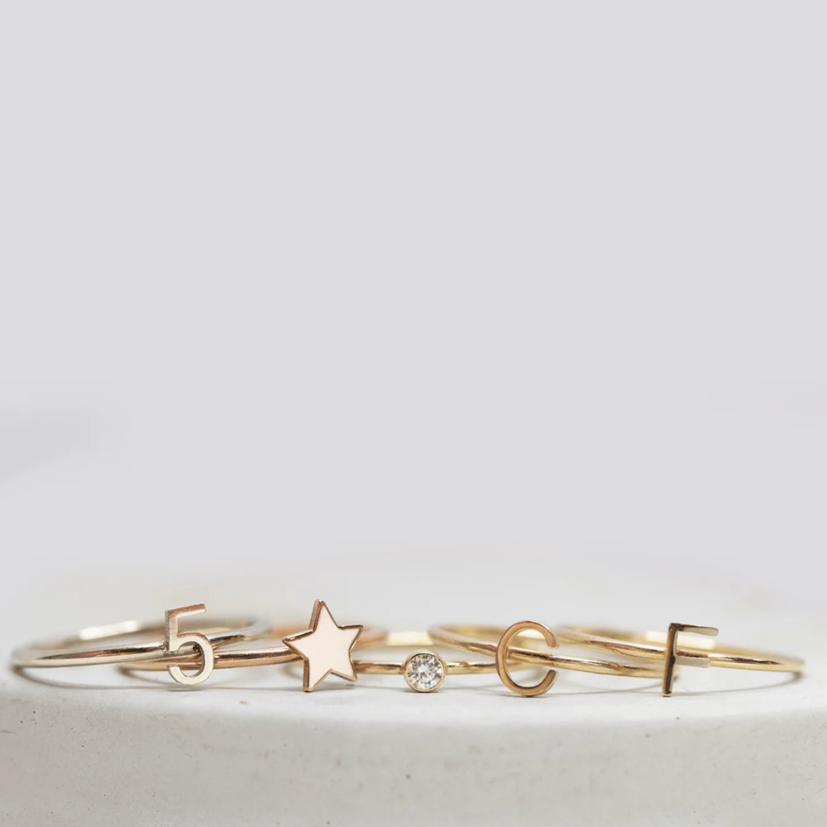 Two 14k gold Letter Rings (C and F), a Lucky Number Ring (5), a Twinkle Twinkle Ring and a Birthstone Ring, against a light grey background.