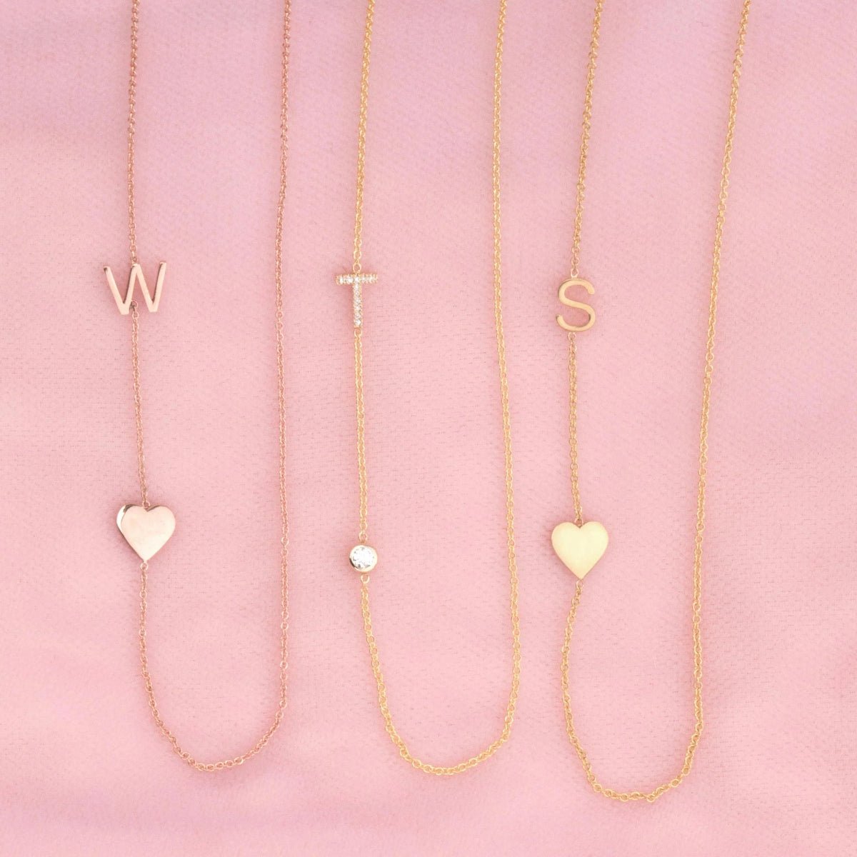 Three necklaces against a pink background -  Monogram Necklace with Heart Rose Gold and a W letter charm, a  Pavé Monogram Necklace with Diamond and a T letter charm, and a Monogram Necklace with Heart Yellow Gold and an S letter charm. 