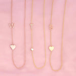 Three necklaces against a pink background -  Monogram Necklace with Heart Rose Gold and a W letter charm, a  Pavé Monogram Necklace with Diamond and a T letter charm, and a Monogram Necklace with Heart Yellow Gold and an S letter charm. 