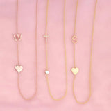 Three necklaces against a pink background -  Monogram Necklace with Heart Rose Gold and a W letter charm, a  Pavé Monogram Necklace with Diamond and a T letter charm, and a Monogram Necklace with Heart Yellow Gold and an S letter charm. 
