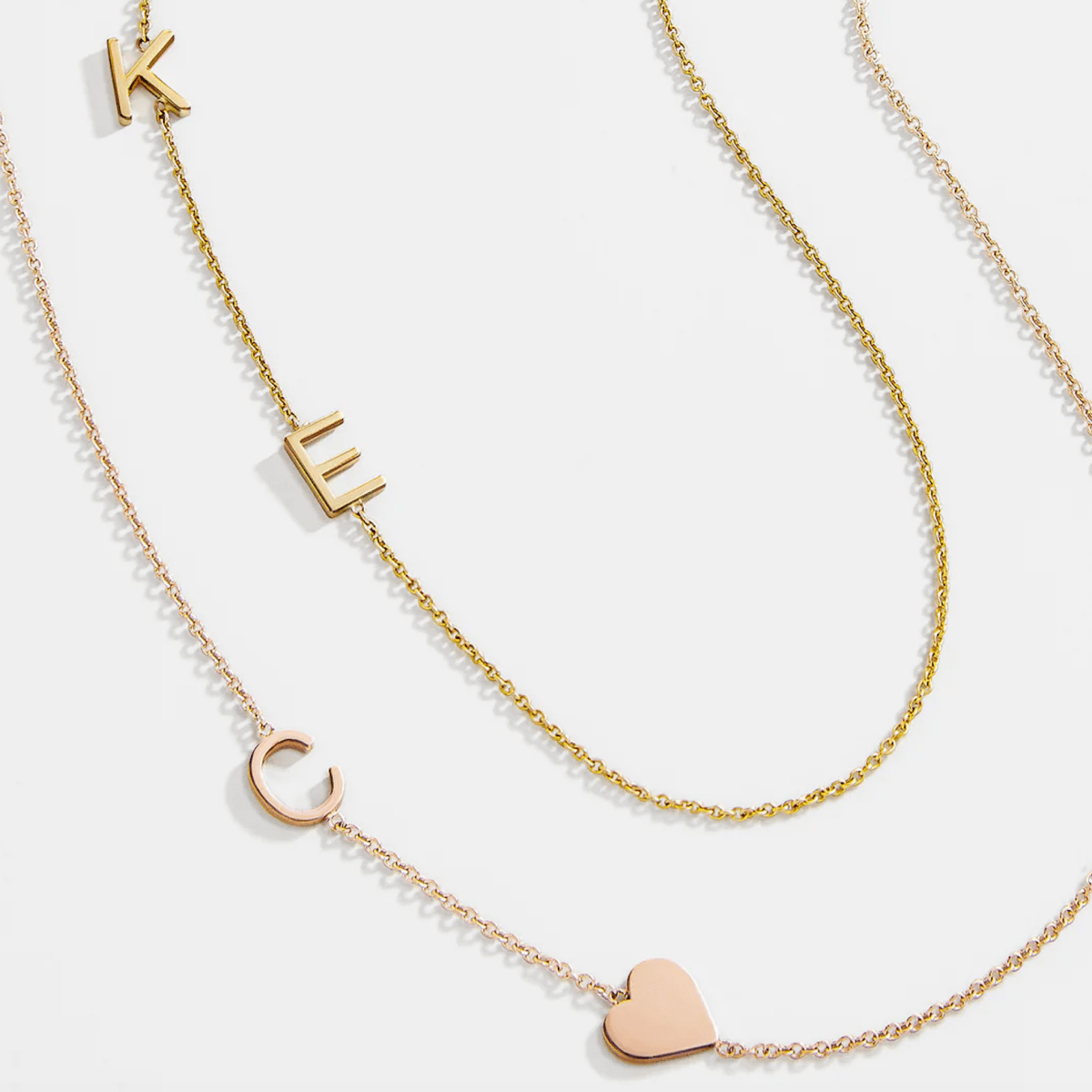 A yellow gold Custom Gold Necklace with two letters (K and E) and a Monogram Necklace with Heart Rose Gold with a letter C charm, against a white background.
