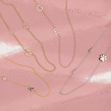 Four Custom Gold Necklaces: a yellow gold necklace with a K letter and birthstone charm, a yellow gold beaded necklace with pavé letter charms J, M and C, a rose gold necklace with two letter charms G and E, and a white gold necklace with two L letter charms and a paw, on a pink background.