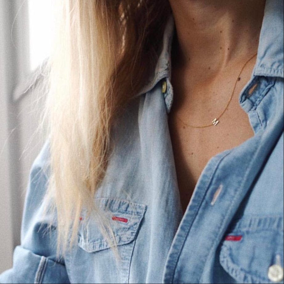 A close-up of a woman's neck and chest area wearing a gold necklace with a letter M charm and a denim shirt.
