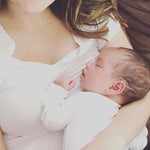 A woman holding a baby and wearing a gold necklace with a B letter charm. 