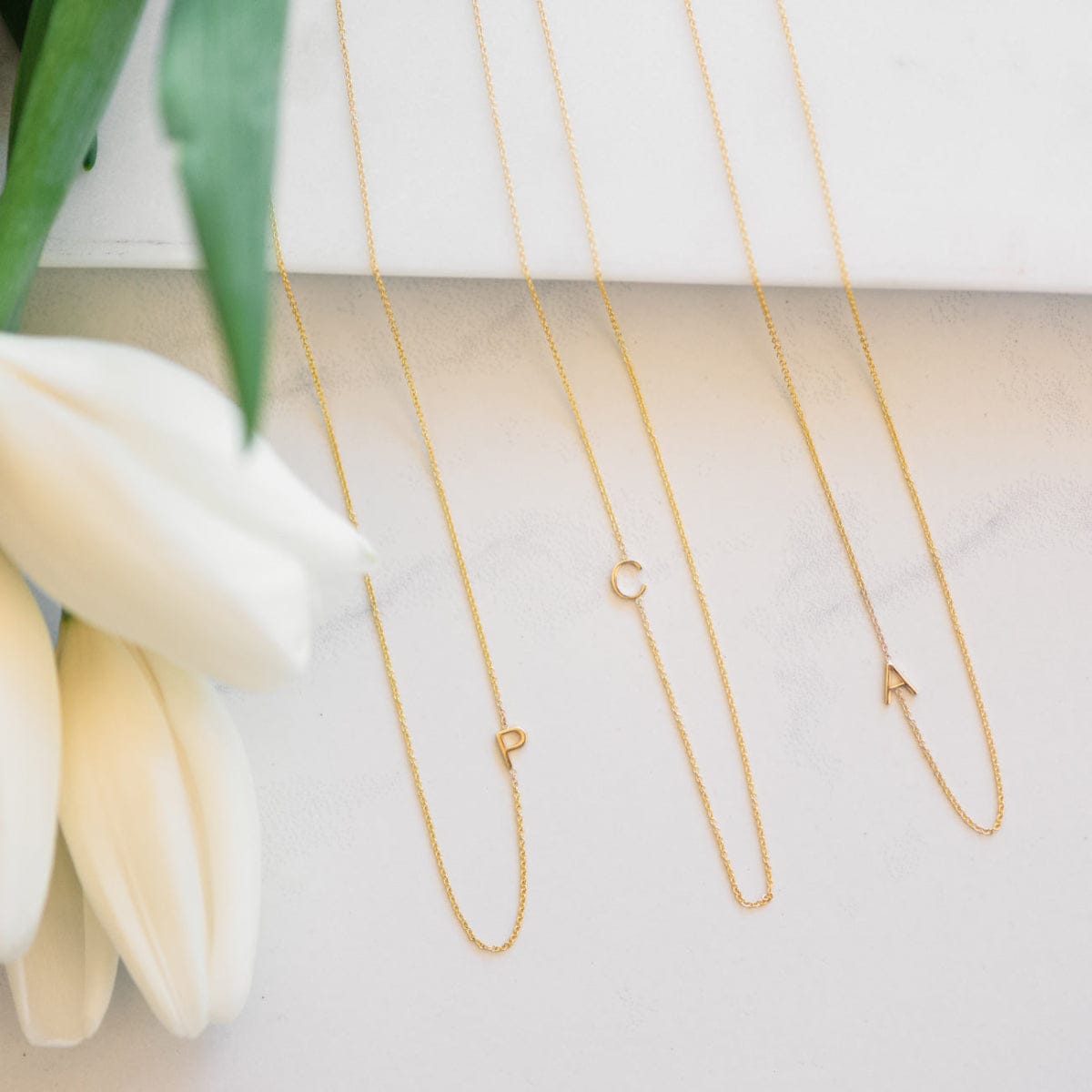 Three gold necklaces with initial charms on a white surface with flowers.