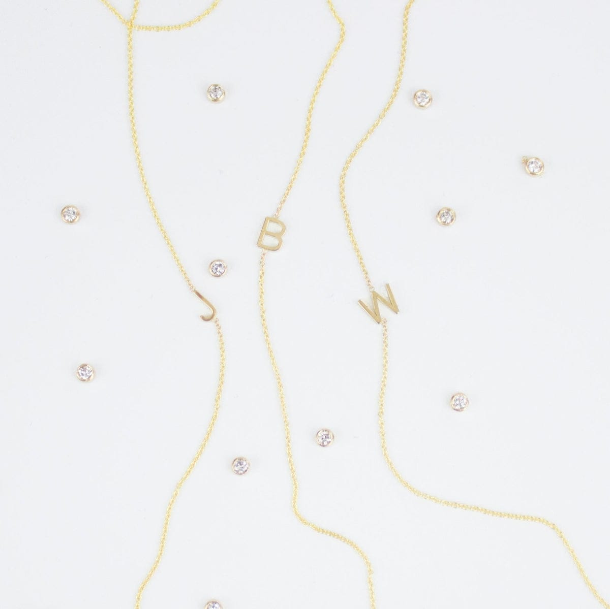 Three gold necklaces with single letter charms, being J, B and W, featuring multiple little gemstones on a white background.