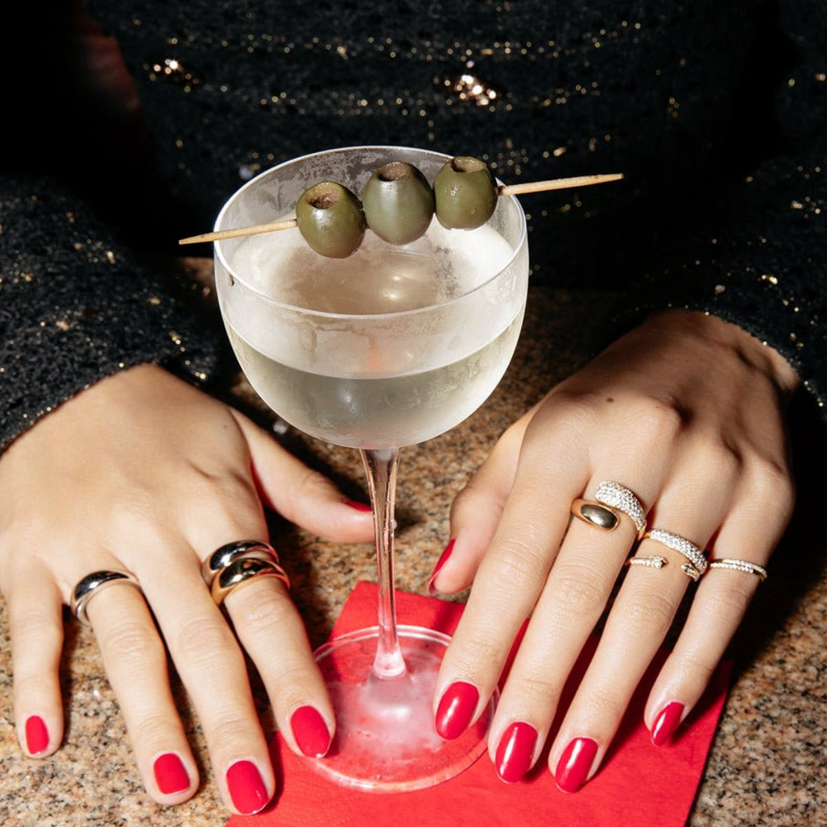 A pair of hands wearing several Martini Rings, a Make it a Double Ring, a Cosmo Ring, an Open Diamond Ring and two Pave Diamond Rings, with red nails holding a martini cocktail. 