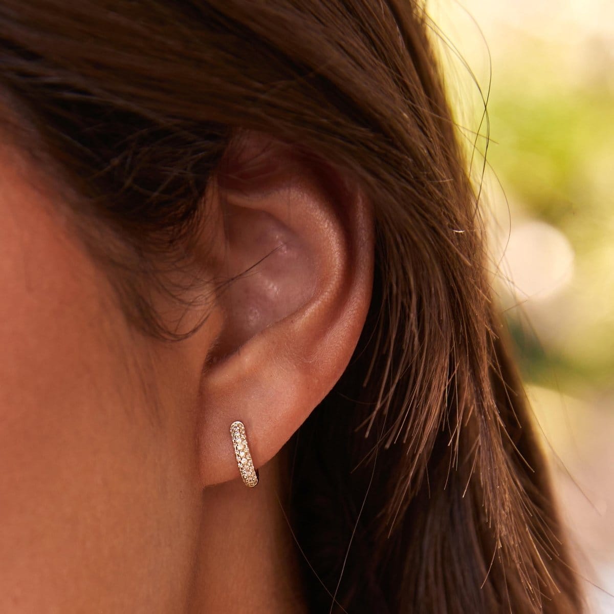 Close-up of Pavé Huggie in a brunette woman's ear with a blurred light green background. 