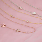 Three necklaces laid on a pink fabric background: a Monogram Necklace with Heart Rose Gold and a letter W charm, a Pavé Monogram Necklace with Diamond and a letter T charm, and a Monogram Necklace with Heart White Gold and a letter S charm.