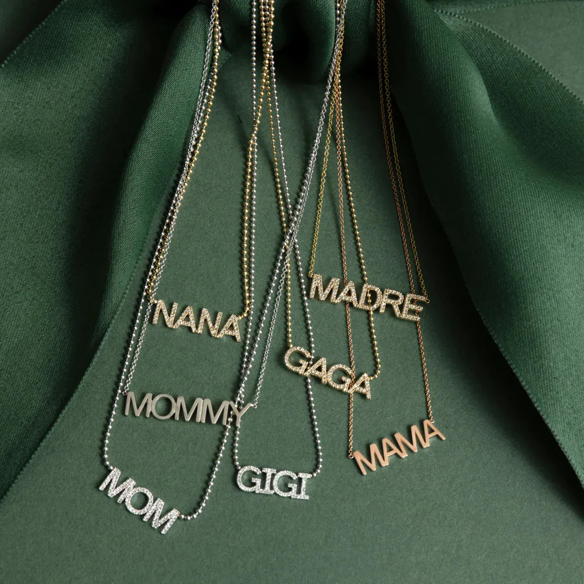 A Pavé NANA Necklace, a MOMMY Necklace, a Pavé MOM Necklace, a Pavé MADRE Necklace, a Pavé GAGA Necklace, Pavé GIGI Necklace and a MAMA Necklace, against a dark green fabric background. 