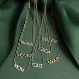 A Pavé NANA Necklace, a MOMMY Necklace, a Pavé MOM Necklace, a Pavé MADRE Necklace, a Pavé GAGA Necklace, Pavé GIGI Necklace and a MAMA Necklace, against a dark green fabric background. 