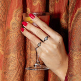 A hand wearing three Pinky Promise Rings with red nails, holding a cocktail against a colourful patterned fabric background. 