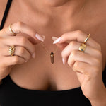 Close-up of a woman's décolletage wearing the Courage Retreat Necklace, with her hands holding the necklace and wearing an Etched Paris Ring, a Thick Band, a Geo Stacking Ring Square, and the Evermore Ring, styled in a black singlet top.