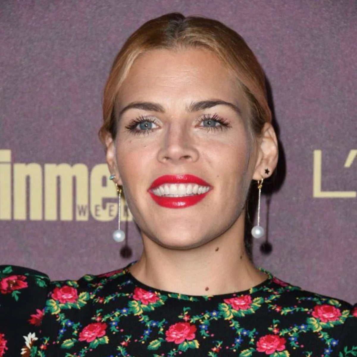 Busy Philipps wearing a 14K Gold Charm Stud - Star, drop pearl earrings, and a black dress with red flowers against a dark purple background.