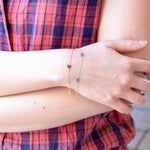 Close-up of a person wearing a plaid shirt and two bracelets - a gold bracelet with a heart charm and a white gold bracelet with two E charms.