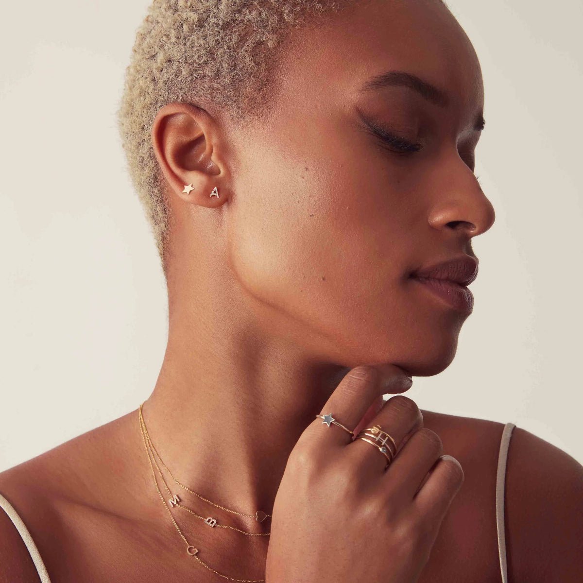 Close-up of a woman wearing two stud earrings, being a star shape and the letter 'A', rings and necklaces with a neutral background. 