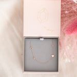 A Custom Gold Necklace in rose gold with two letters, G and K on the left side and an A and a heart on the right side, in a Maya Brenner jewellery box against a neutral background with pink feathers.