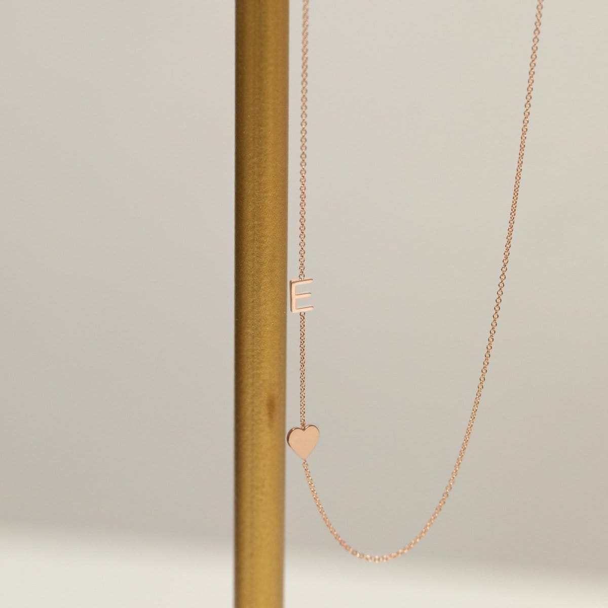 A Monogram Necklace with Heart Rose Gold and a letter E charm next to a brown stick against a light grey background. 