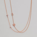 A Pavé Monogram Necklace with Diamond and a letter T charm in rose gold, and a Monogram Necklace with Heart Rose Gold and a letter S charm, against a neutral background.