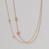 A Pavé Monogram Necklace with Diamond and a letter T charm in rose gold, and a Monogram Necklace with Heart Rose Gold and a letter S charm, against a neutral background.