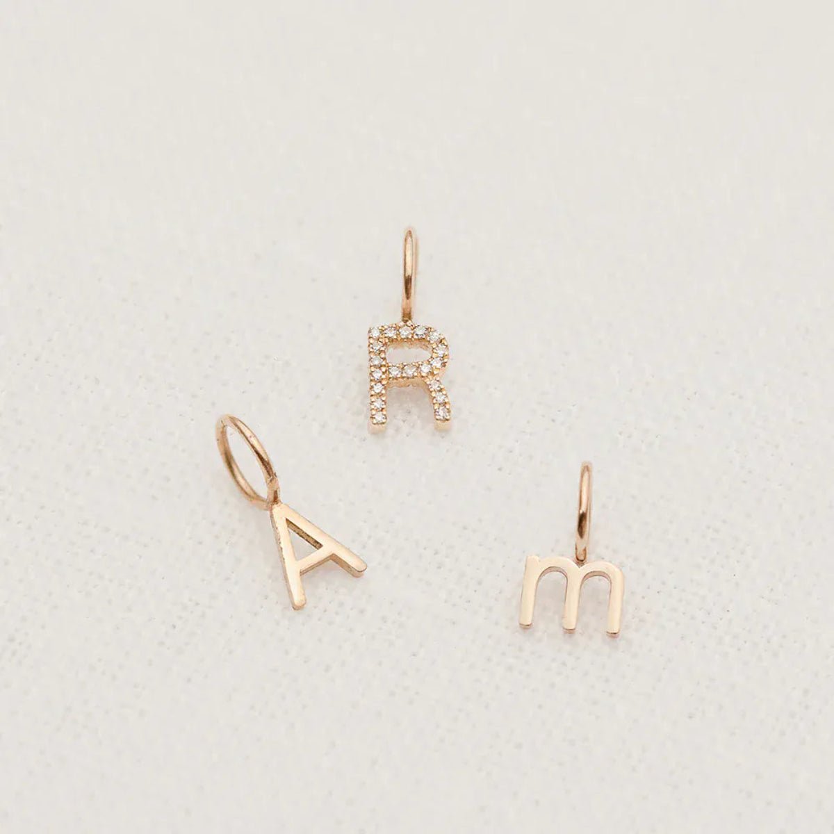A Petite Letter Charm in m, a Gold Letter Charm in A and a Pavé Letter Charm in R, all in rose gold, against a white textured background. 