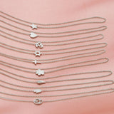 Nine white gold 14k Gold Asymmetrical Charm Necklaces with cable chains (star, heart, om, star of david, cross, paw, angel wing, hamsa and recovery) laid vertically on a light pink fabric background.