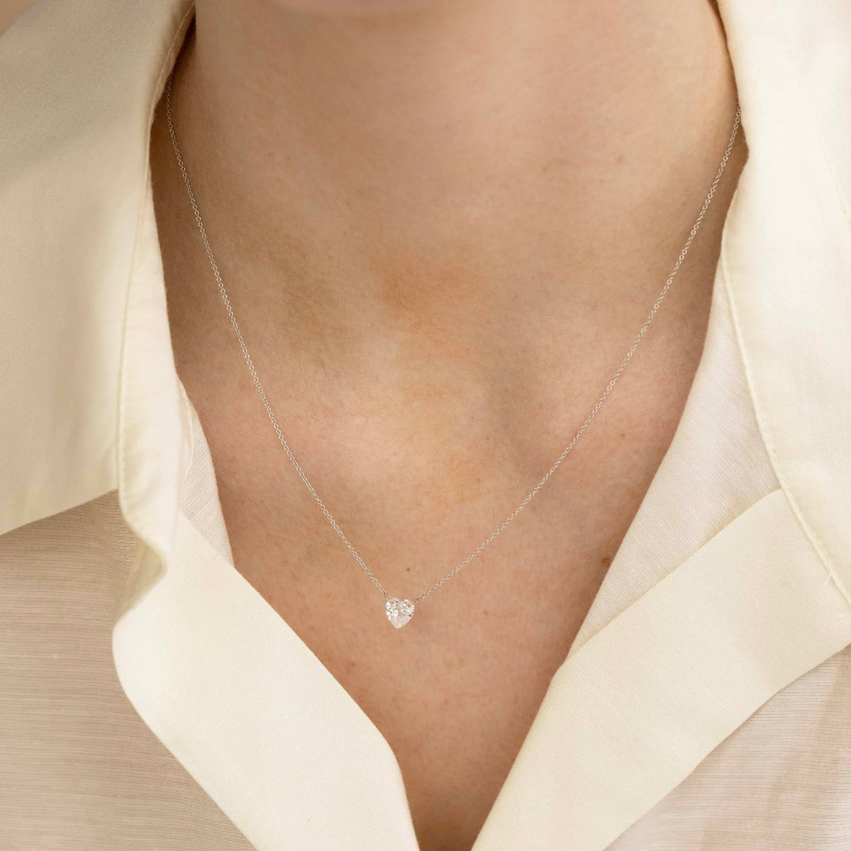 Close-up of a décolletage wearing The Sweetheart Necklace - White Sapphire in white gold with a cream shirt.