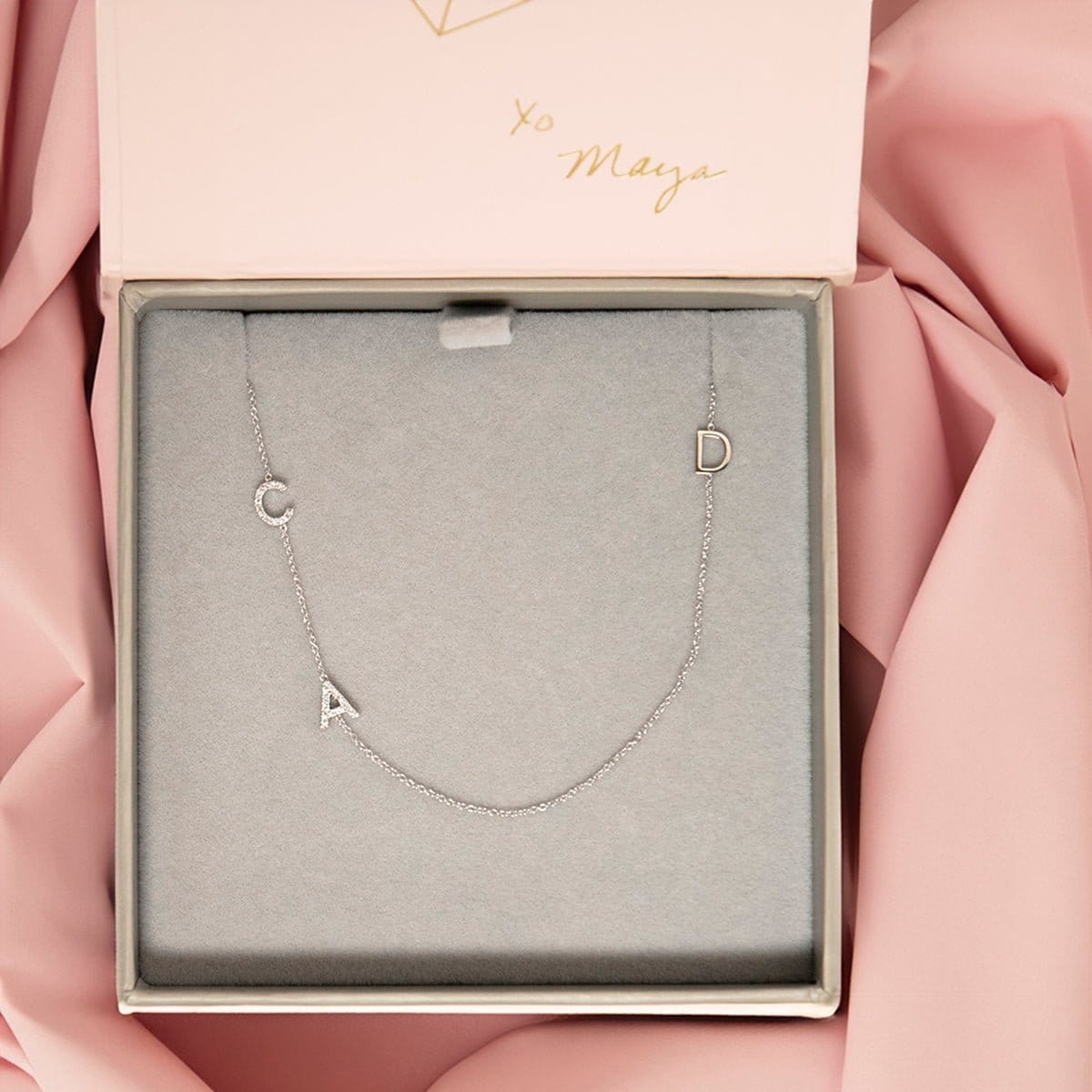 A Custom Gold Necklace with three letters, a pavé C and A on the left side and a D on the right side, in a Maya Brenner case against a pink fabric background.