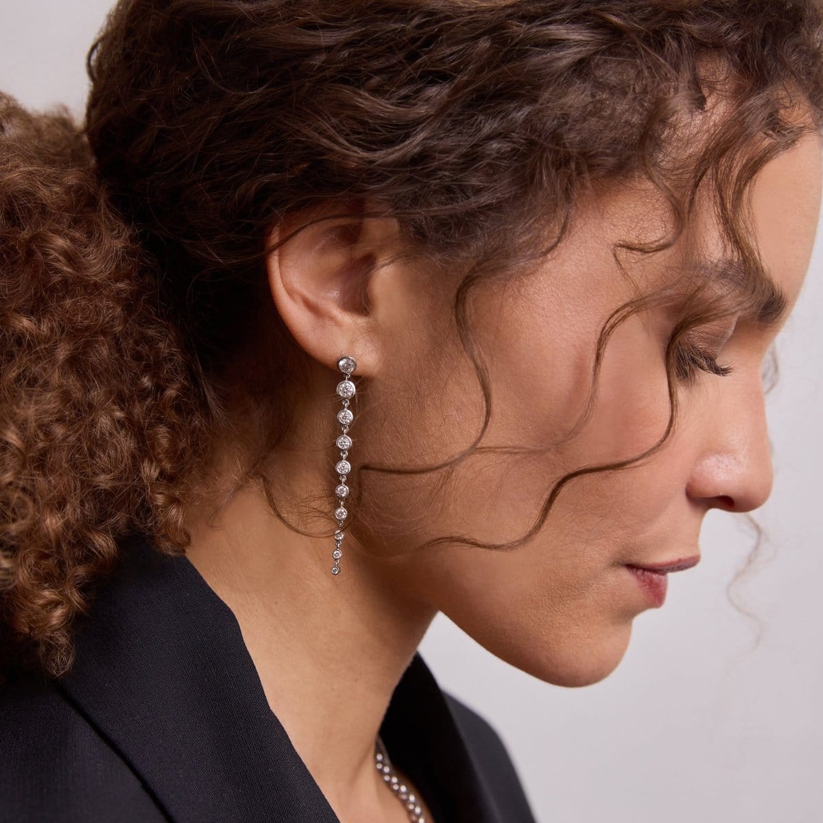 Close-up of a woman's side profile wearing a white gold Diamond Cascade Earring, with brown curly hair and a black top on a neutral background. 