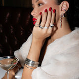 A woman wearing the Evermore Ring, a Martini Ring, a Cosmo Ring, a Make It a Double Ring, and a Love on the Rocks Ring, along with the Triple Sec Diamond Earring, the Night Cap Ear Cuff, a Diamond Cascade Earring and several For Keeps Bangles, all in white gold, with red nails and red lipstick, dressed in a cream jacket and holding a cocktail against a dark background.